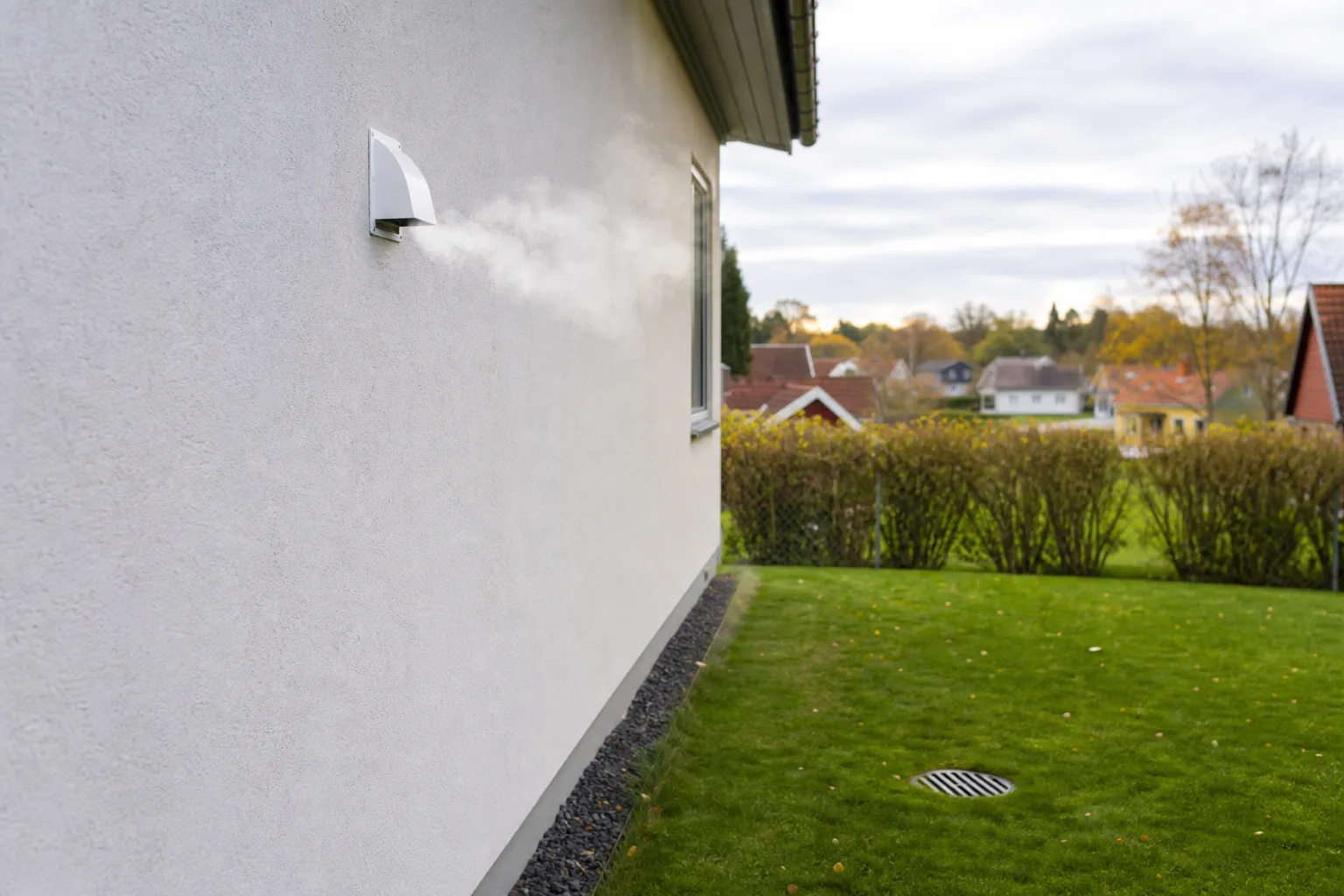 En vit ventilationshuv monterad högt på en putsad svensk villafasad, med gräsmatta och en dagvattenbrunn synlig längre ner